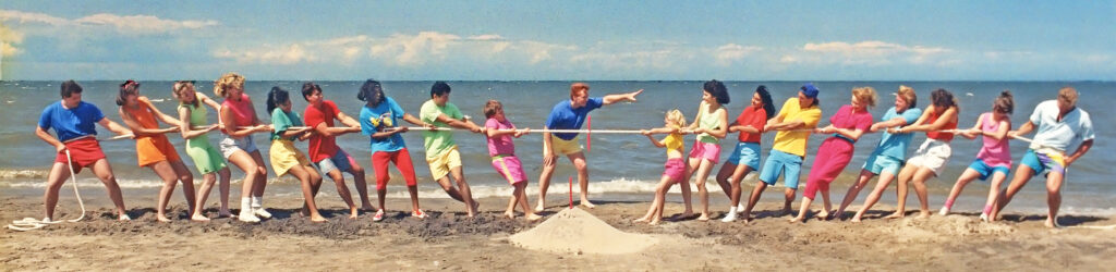 A group of people play tug of war on a beach. A referee stands in the middle.